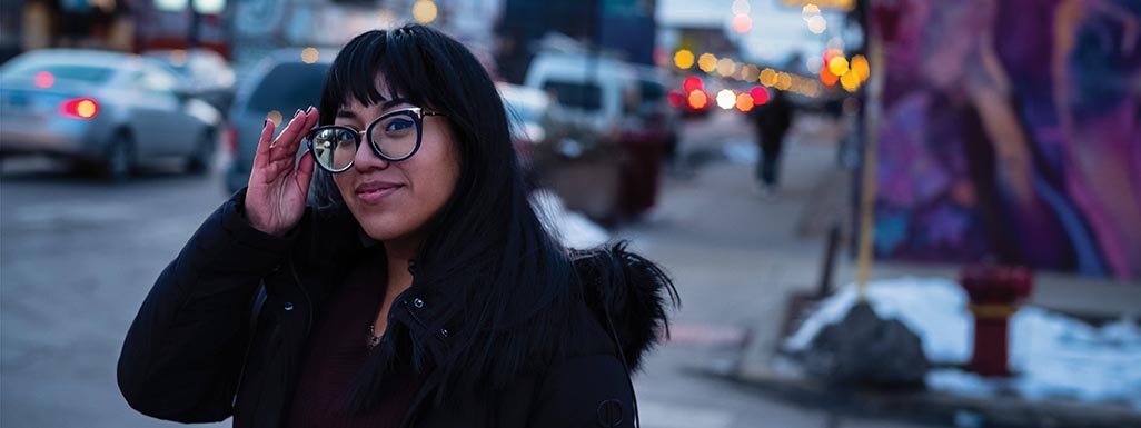 A smiling woman posing on an urban street corner with artwork, cars, and traffic lights in the background