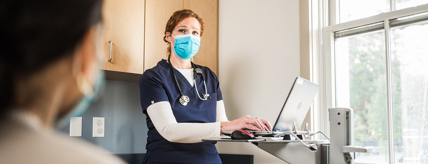 female nurse with a mask on at a laptop with a patient