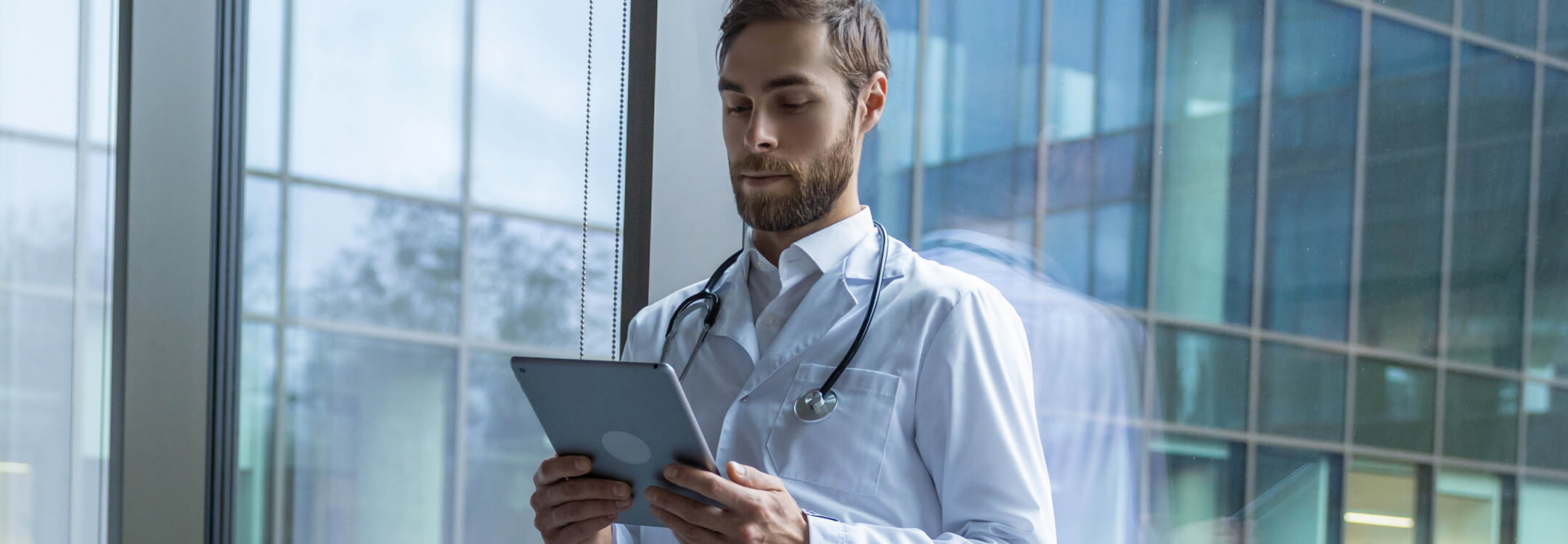 Physician wearing a white coat and stethoscope looks at the electronic health record for a patient on a tablet.