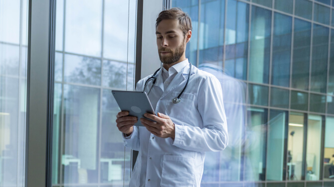 Physician wearing a white coat and stethoscope looks at the electronic health record for a patient on a tablet.