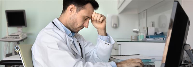 A doctor at a desk, visibly stressed, with his head in his hands, reflecting on healthcare challenges and physician burnout.