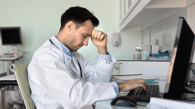 A doctor at a desk, visibly stressed, with his head in his hands, reflecting on healthcare challenges and physician burnout.