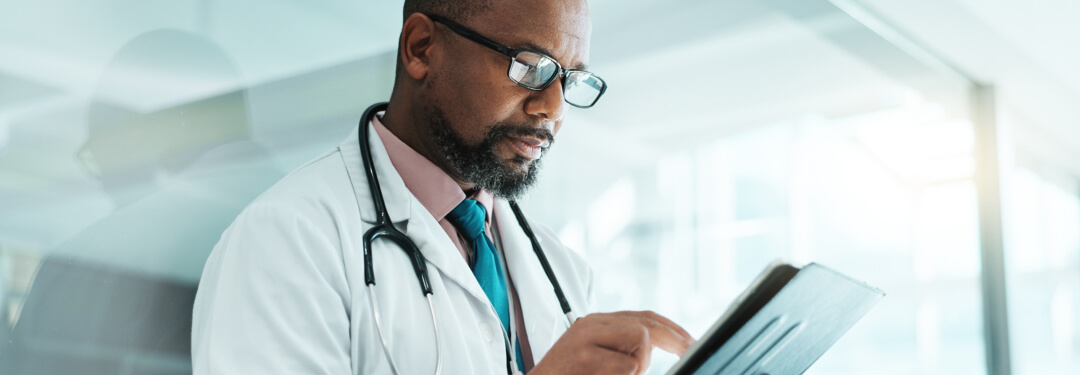A male physician in a white coat reviews data or patient information on a tablet, appearing engaged and attentive.