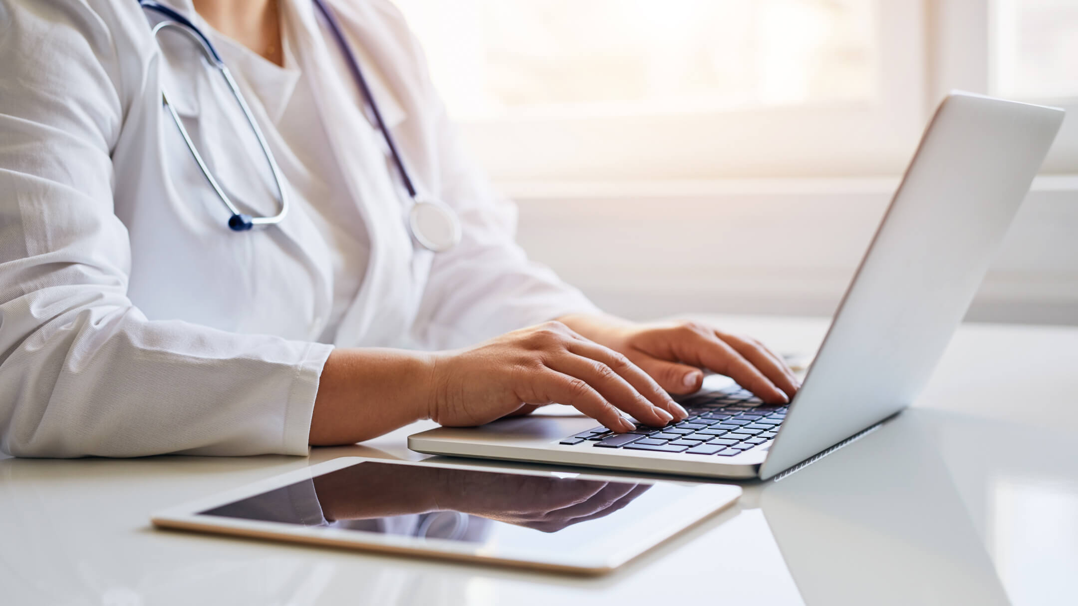 Healthcare professional documenting on a laptop and tablet on desk