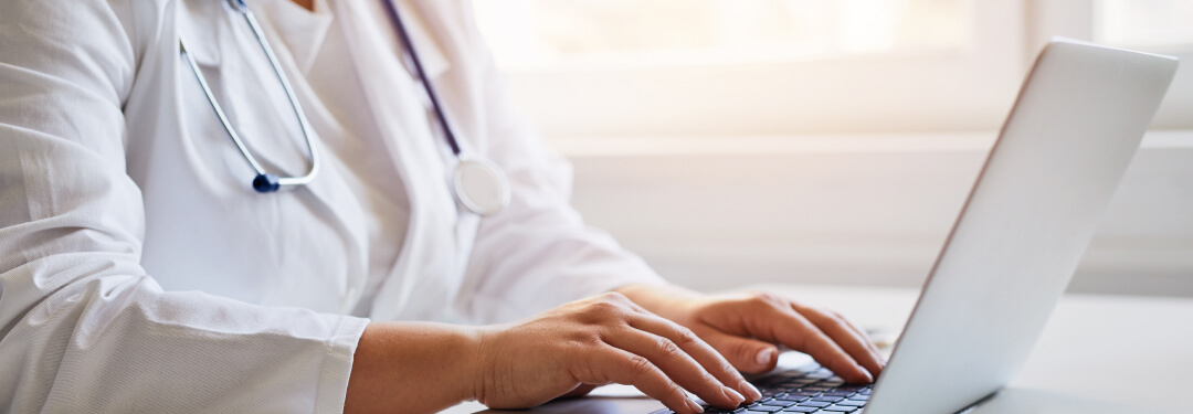 Healthcare professional documenting on a laptop and tablet on desk.