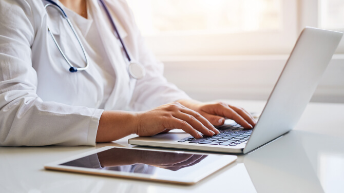 Healthcare professional documenting on a laptop and tablet on desk