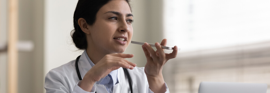A female doctor sitting in front of a laptop utilizes AI-enabled voice navigation on her phone.