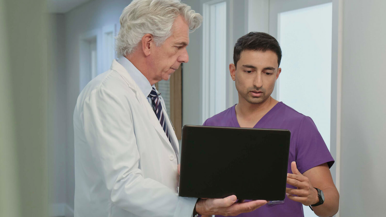 Physician wearing a white coat shows a colleague in scrubs his laptop