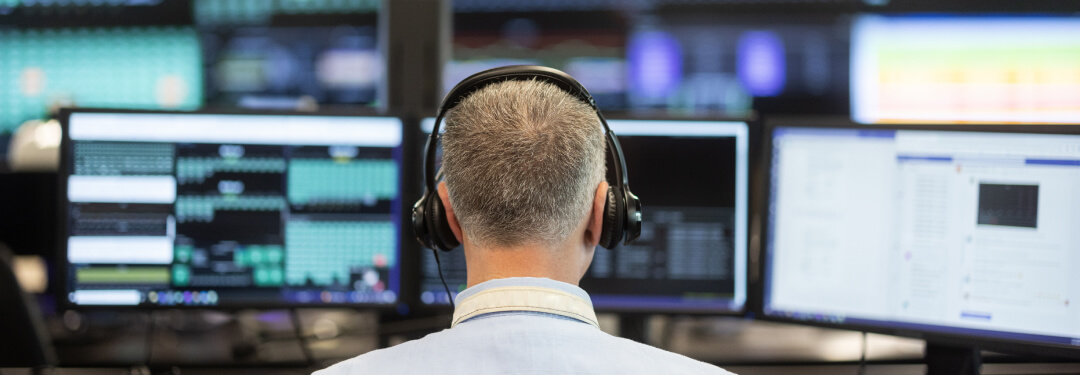 A man wearing headphones works at a desk with several monitors, analyzing information related to healthcare cybersecurity.