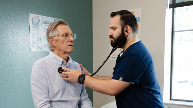 A healthcare professional uses a stethoscope to listen to patient's chest during a medical exam in a clinic room.