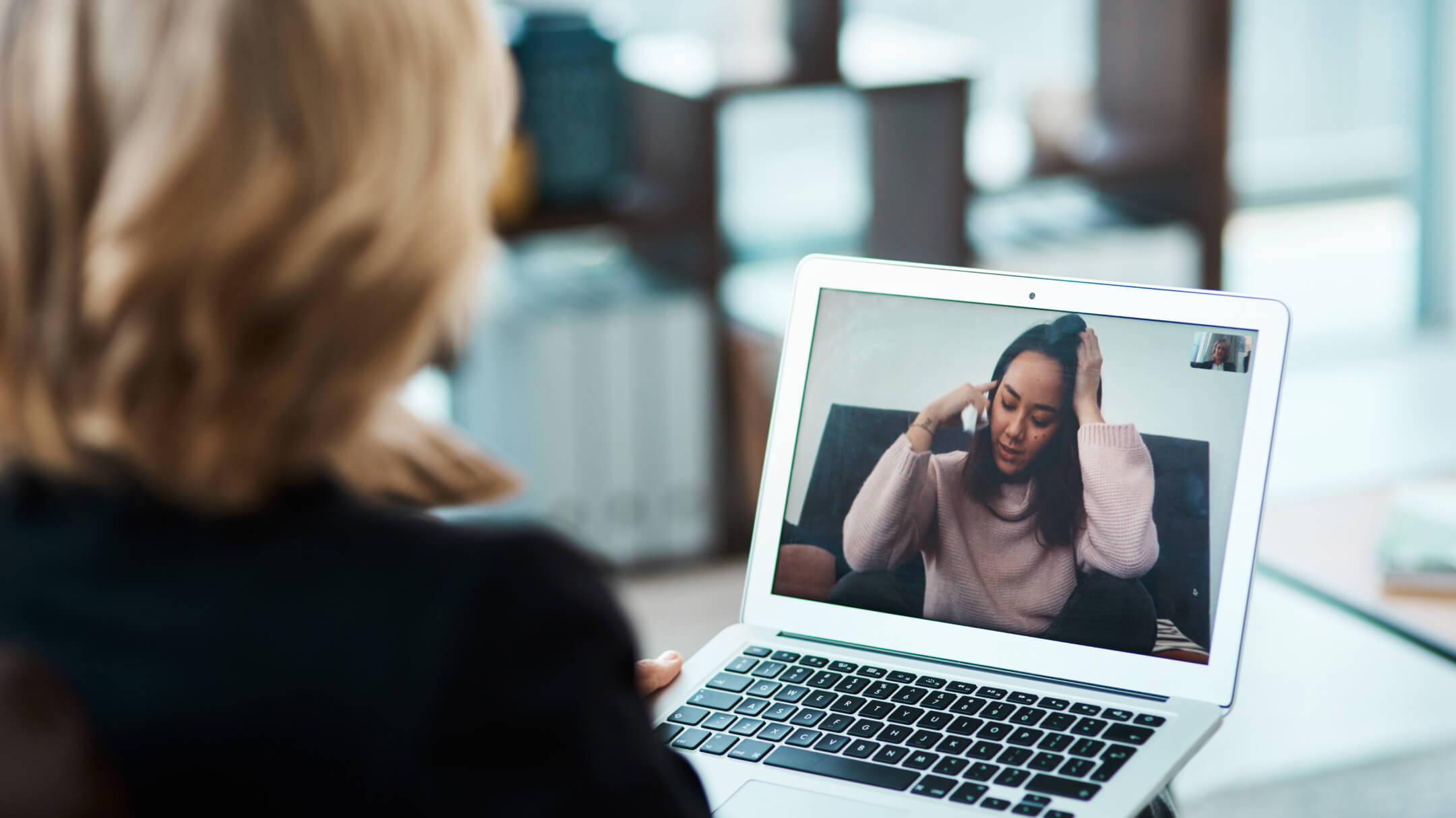 On a woman's laptop screen, a virtual visit is in progress with another woman sitting cross-legged holding her head. 