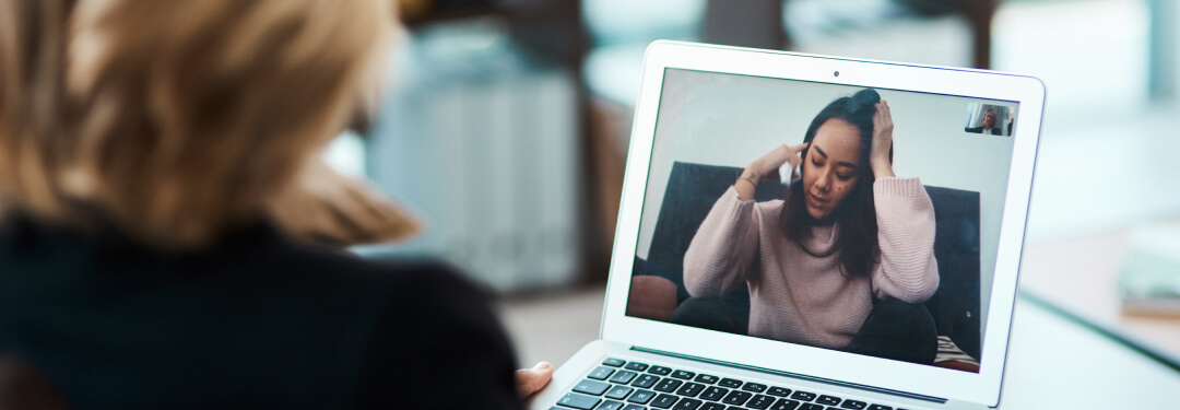 On a woman's laptop screen, a virtual visit is in progress with another woman sitting cross-legged holding her head.