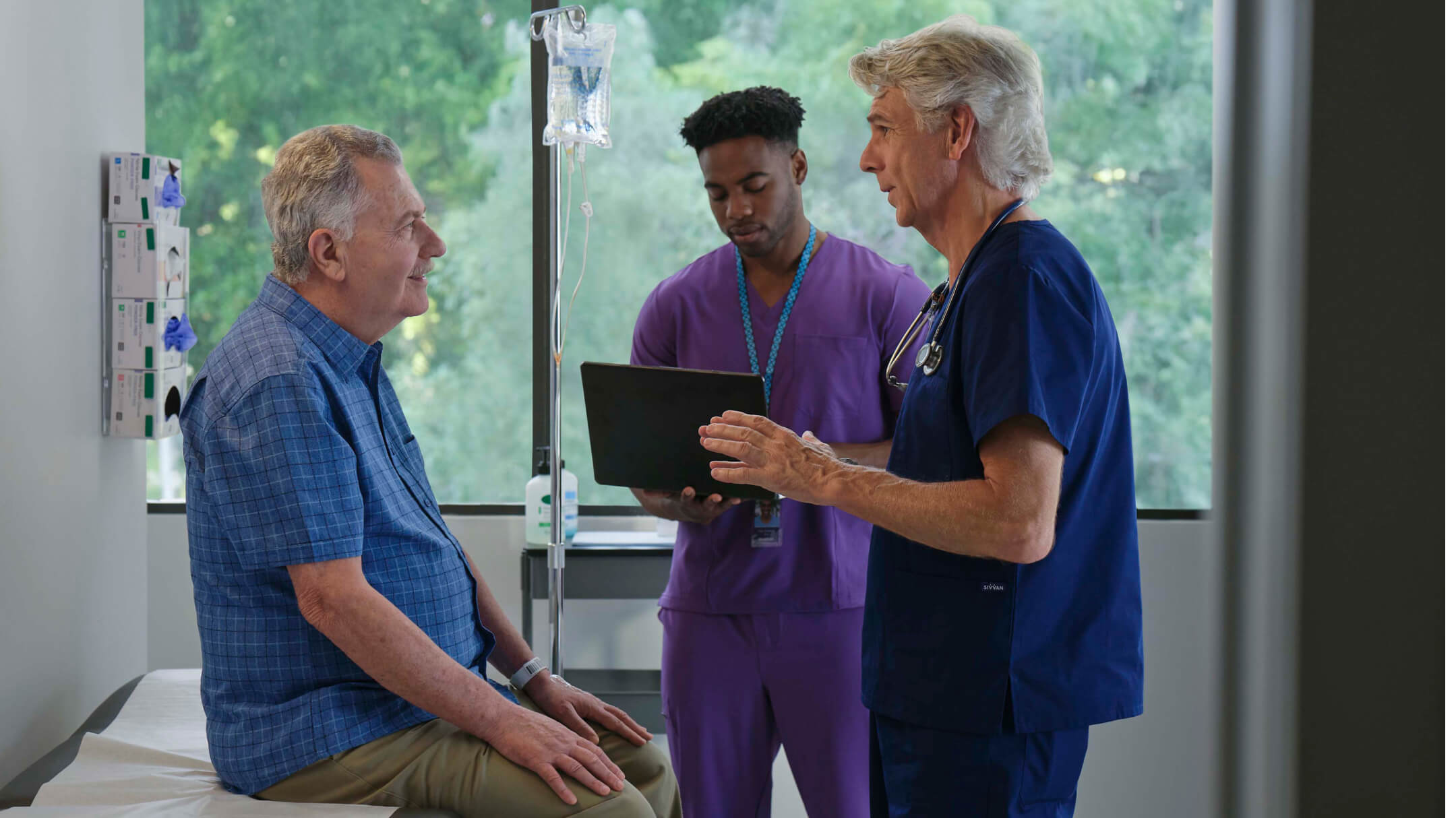 A doctor speaks with a patient while another doctor consults a laptop.