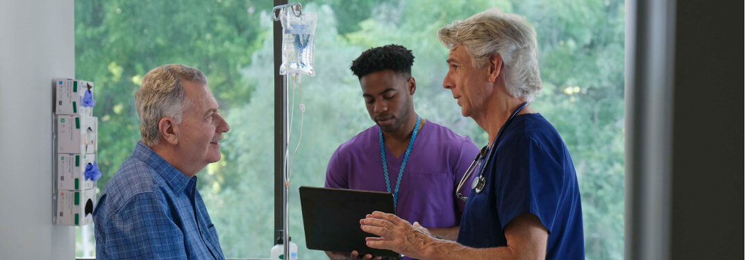 A doctor speaks with a patient while another doctor consults a laptop.