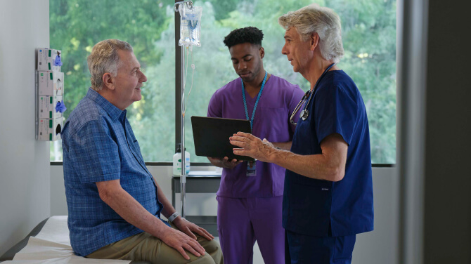 A doctor speaks with a patient while another doctor consults a laptop.