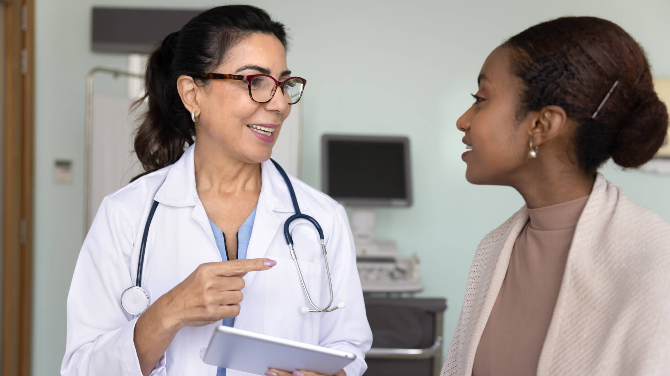 Physician wearing white coat and stethoscope holding a tablet while seeing a patient in medical exam room. 