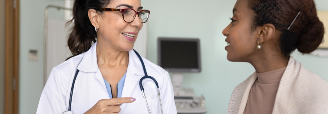 Physician wearing white coat and stethoscope holding a tablet while seeing a patient in medical exam room.