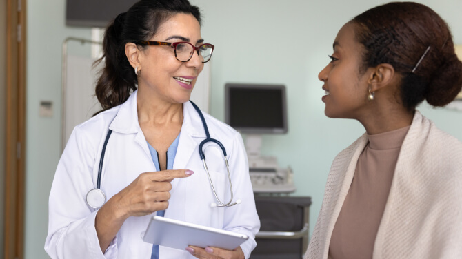 Physician wearing white coat and stethoscope holding a tablet while seeing a patient in medical exam room.
