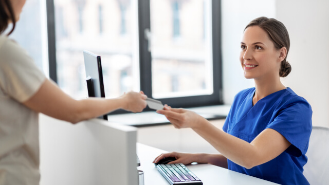 A medical assistant in blue scrubs hands a credit card to the patient, symbolizing improved revenue cycle efficiencies.