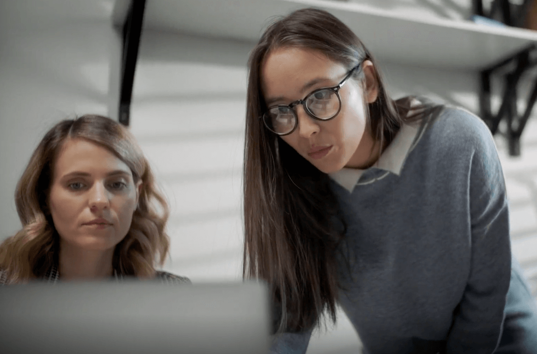 Two women, one standing concentrating on the laptop display in front of them