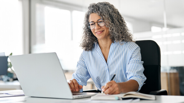 There is a woman with curly hair sitting at a desk working on her laptop while taking notes in a book with a pen.