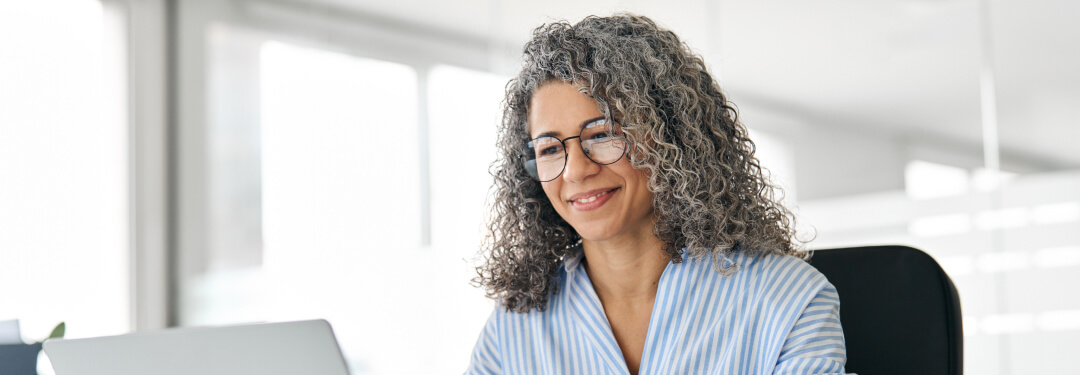 There is a woman with curly hair sitting at a desk working on her laptop while taking notes in a book with a pen.