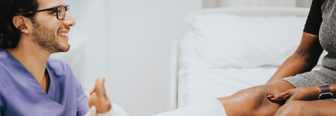 A nurse wearing scrubs smiles as they examine a patient's leg within a healthcare facility.