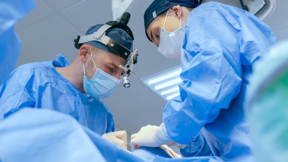Two surgeons in blue scrubs and surgical masks work together in an operating room, focused on a medical procedure.