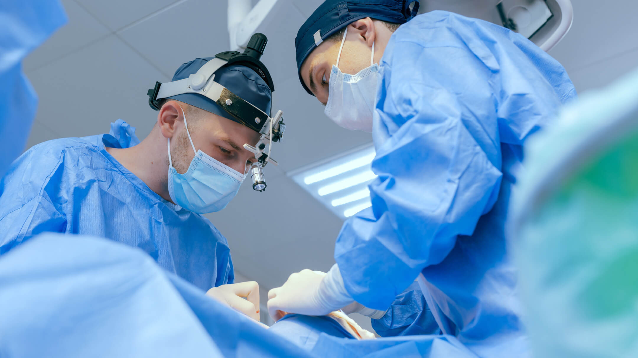 Two surgeons in blue scrubs and surgical masks work together in an operating room, focused on a medical procedure.
