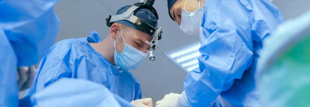 Two surgeons in blue scrubs and surgical masks work together in an operating room, focused on a medical procedure.