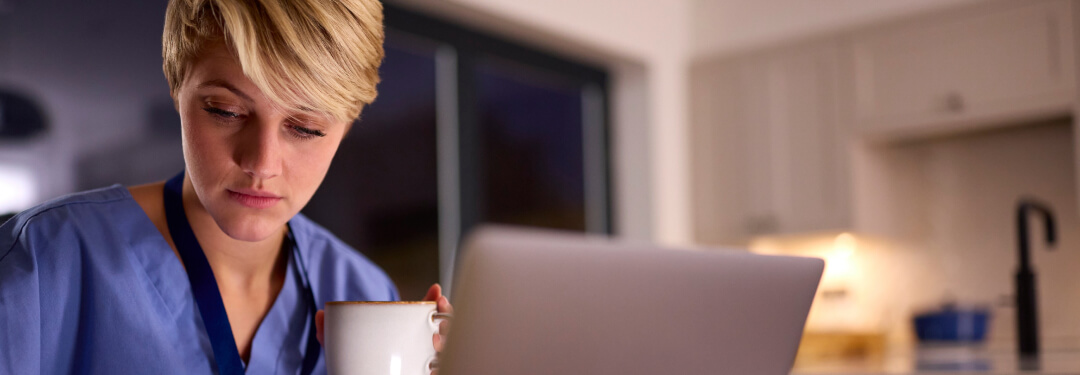 A healthcare professional in scrubs consumes coffee in her office after hours, while taking notes from a laptop.