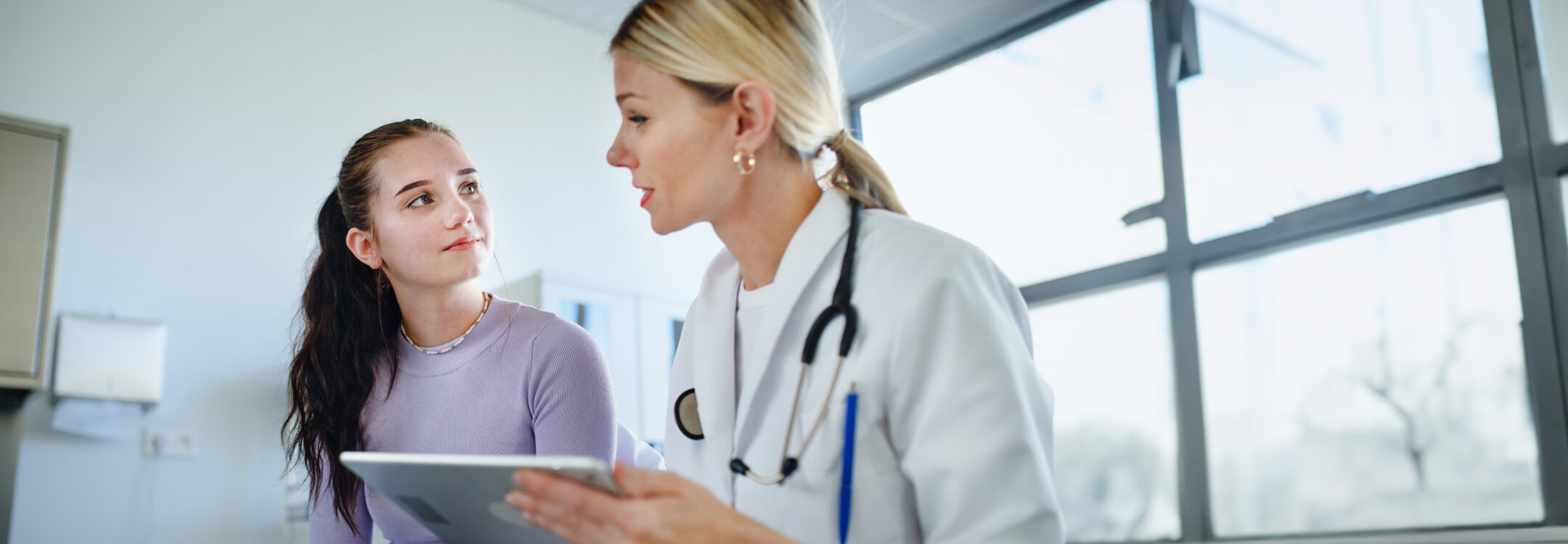 A doctor with a stethoscope discusses with a young patient in a well-lit medical office, unlocking strategic advantages with athenaPayer®.