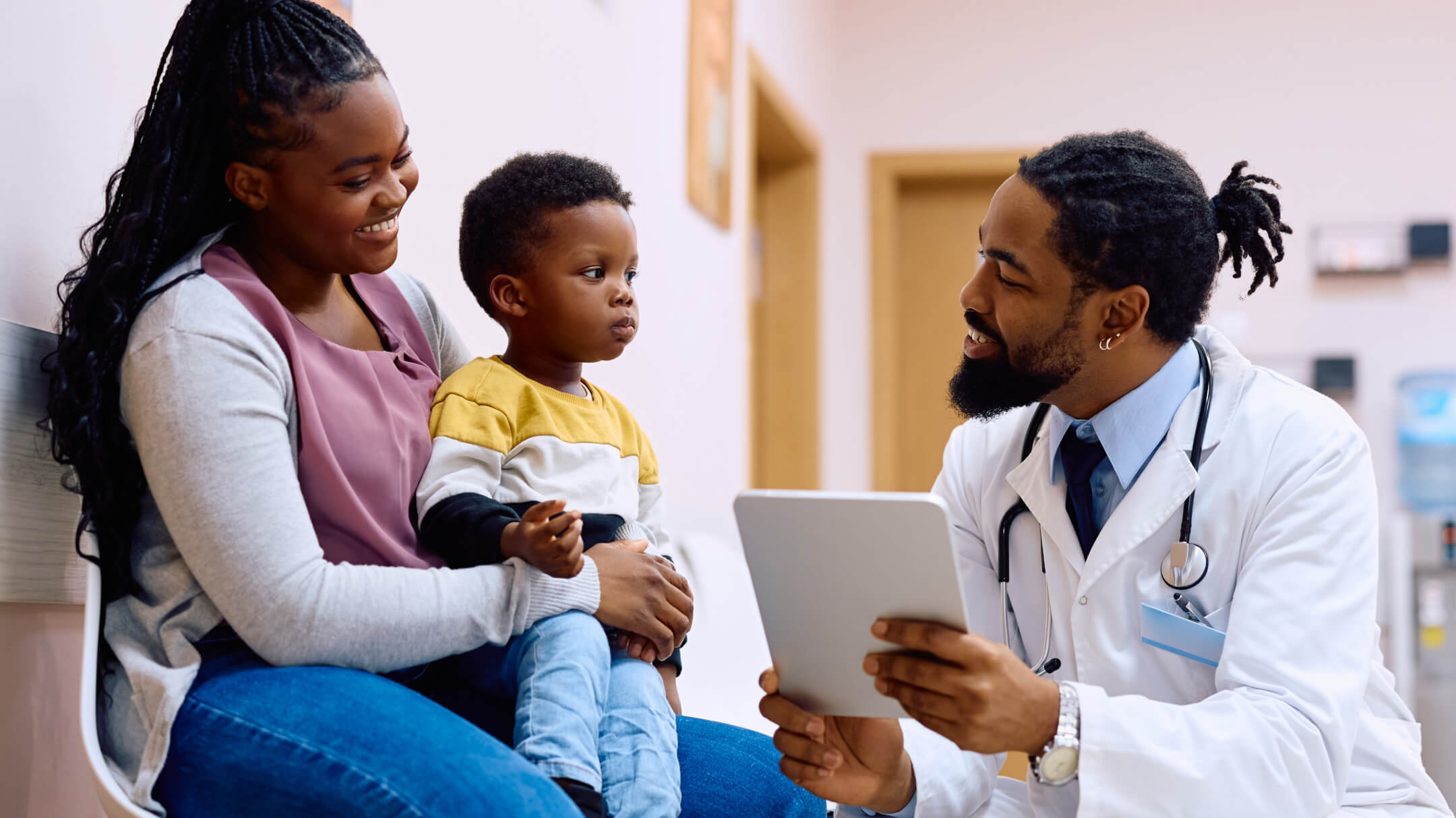 Mother and child speaking with a pediatrician kneeling in front of them holding a tablet.