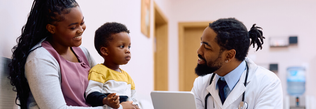 Mother and child speaking with a pediatrician kneeling in front of them holding a tablet.