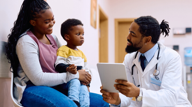 Mother and child speaking with a pediatrician kneeling in front of them holding a tablet.
