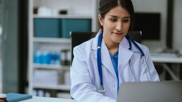 A physician sits at their desk and uses their laptop to engage with their EHR.