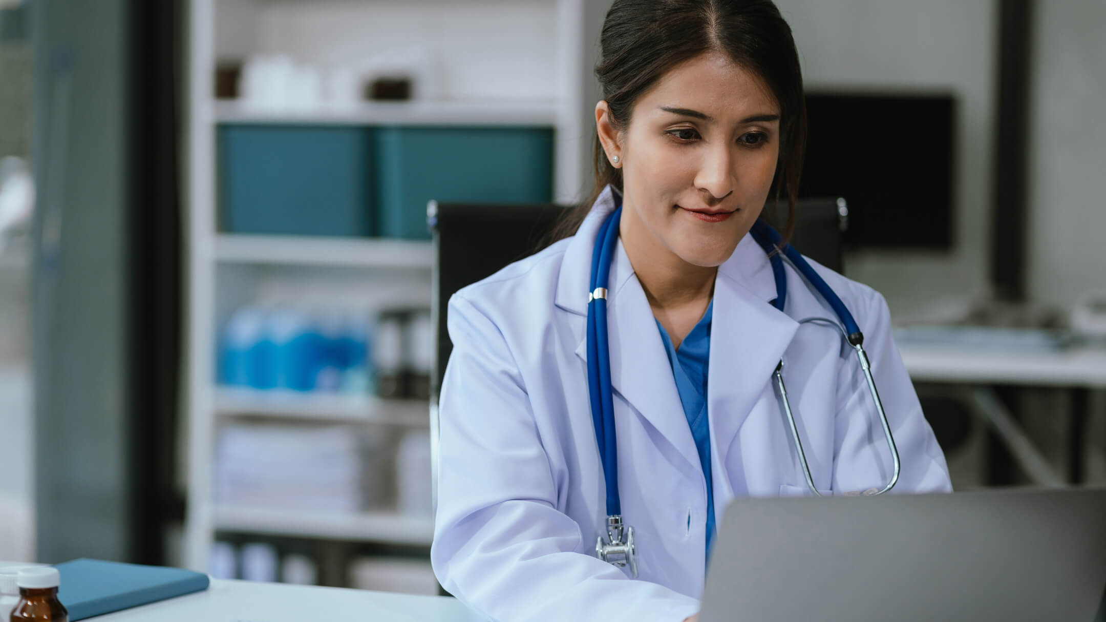 A physician sits at their desk and uses their laptop to engage with their EHR.