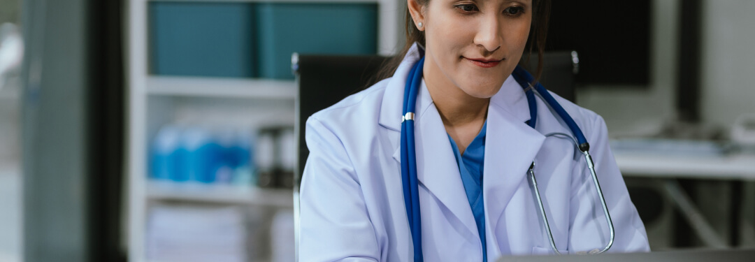 A physician sits at their desk and uses their laptop to engage with their EHR.