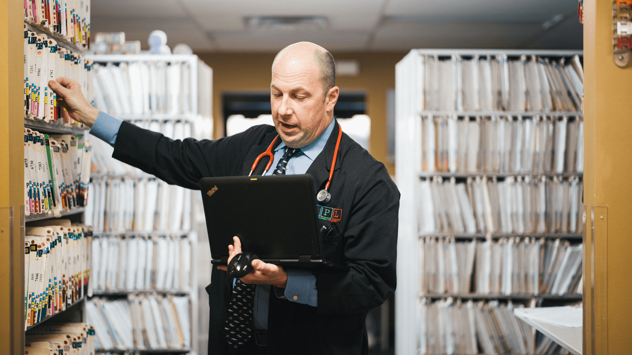 Healthcare professional using a laptop in a medical records room with organized files, illustrating athenaPayer® as a smarter alternative to chart chasing.